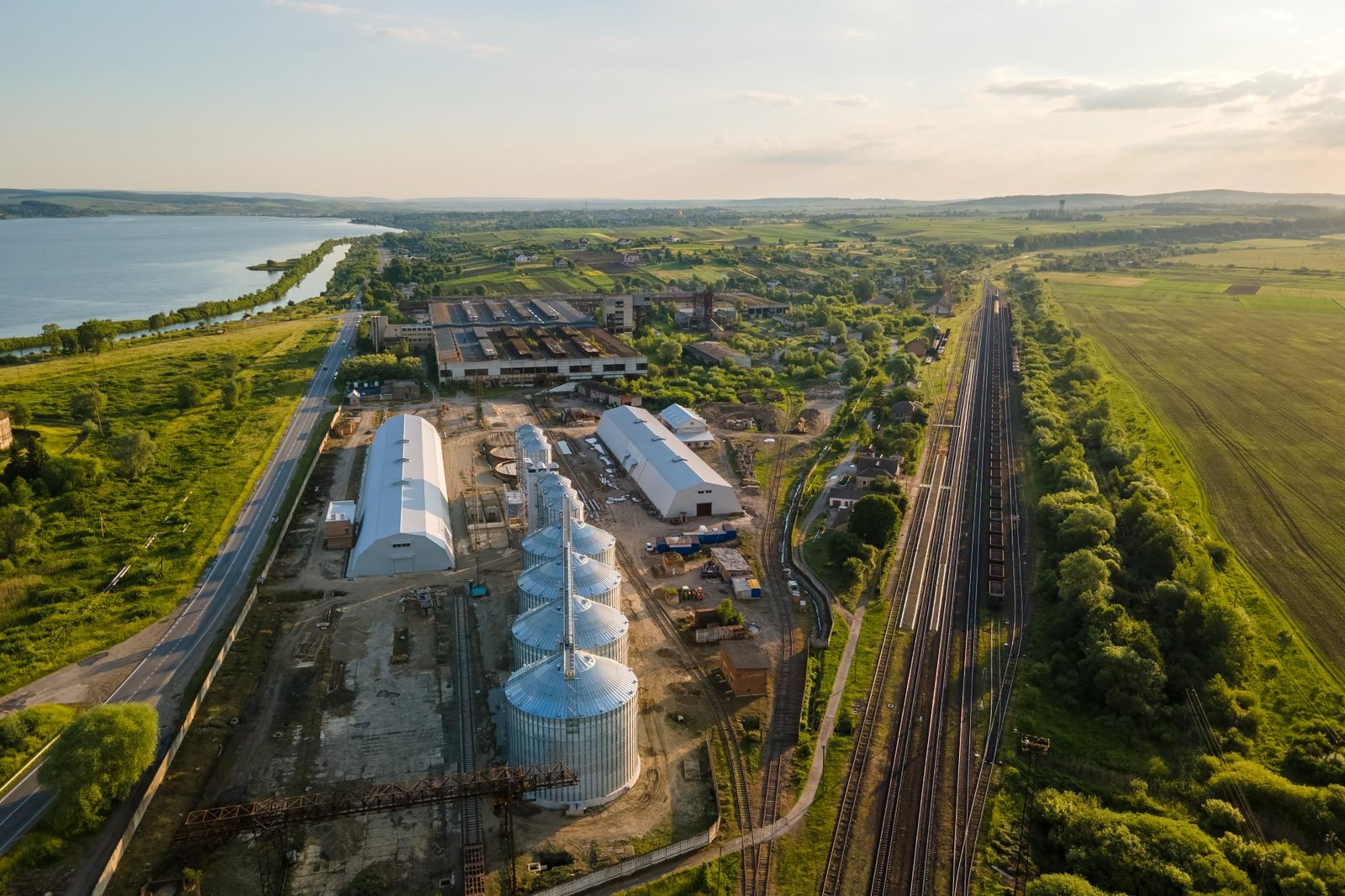 Vista aérea de un polígono industrial junto a una vía férrea y una zona agrícola, con naves, silos metálicos y un entorno verde próximo a un embalse.”